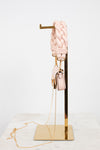 A light pink braided leather shoulder strap with gold-tone hardware, displayed on a gold metal stand next to a matching small pink handbag with a chain strap, set against a clean white background.