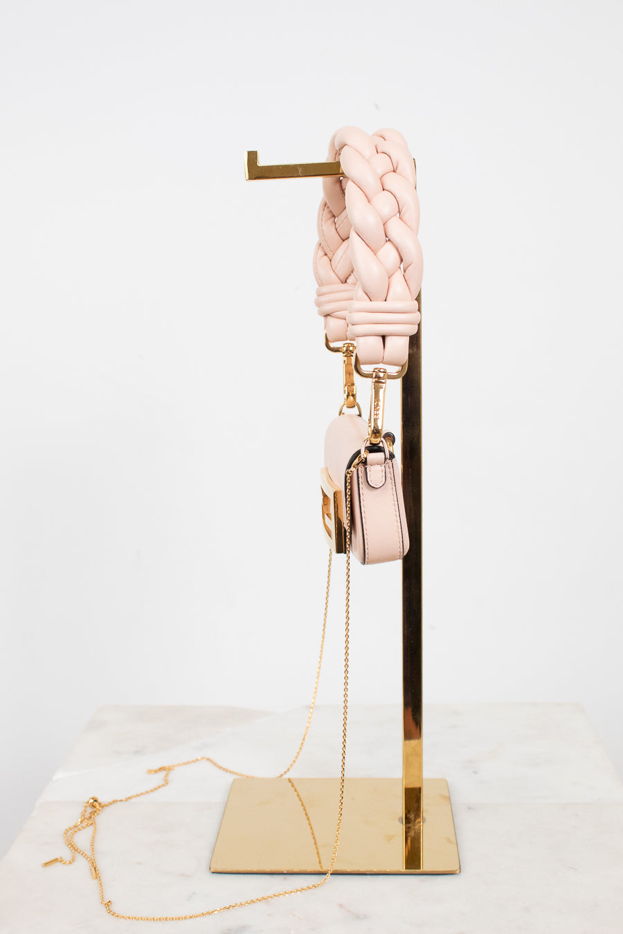 A light pink braided leather shoulder strap with gold-tone hardware, displayed on a gold metal stand next to a matching small pink handbag with a chain strap, set against a clean white background.