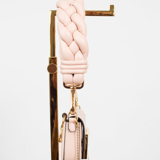 A light pink braided leather shoulder strap with gold-tone hardware, hanging from a gold display stand next to a matching small pink handbag with a gold chain strap, against a plain white background.