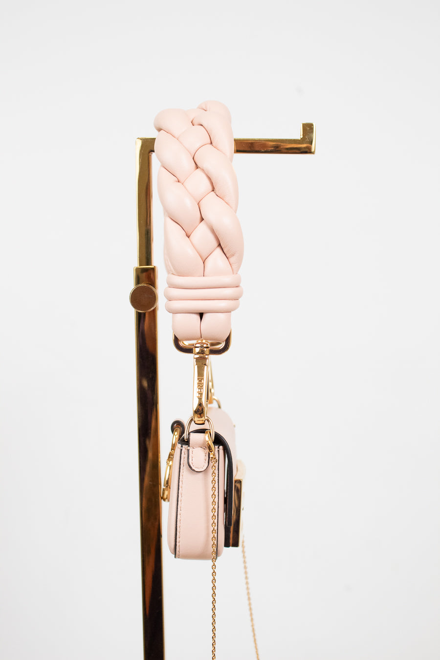 A light pink braided leather shoulder strap with gold-tone hardware, hanging from a gold display stand next to a matching small pink handbag with a gold chain strap, against a plain white background.