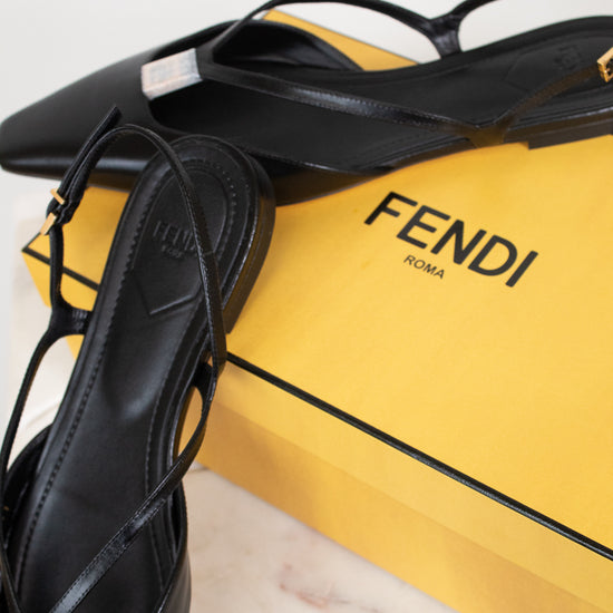 Black Fendi strappy flat shoes displayed next to a bright yellow Fendi box on a light marble surface.