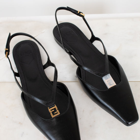 A pair of black leather pointed-toe slingback flats with gold-tone Fendi FF logo hardware, displayed on a light marble surface.