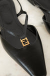 A close-up of a black leather pointed-toe slingback shoe with a gold-tone interlocking FF logo hardware on the strap, resting on a light marble surface.