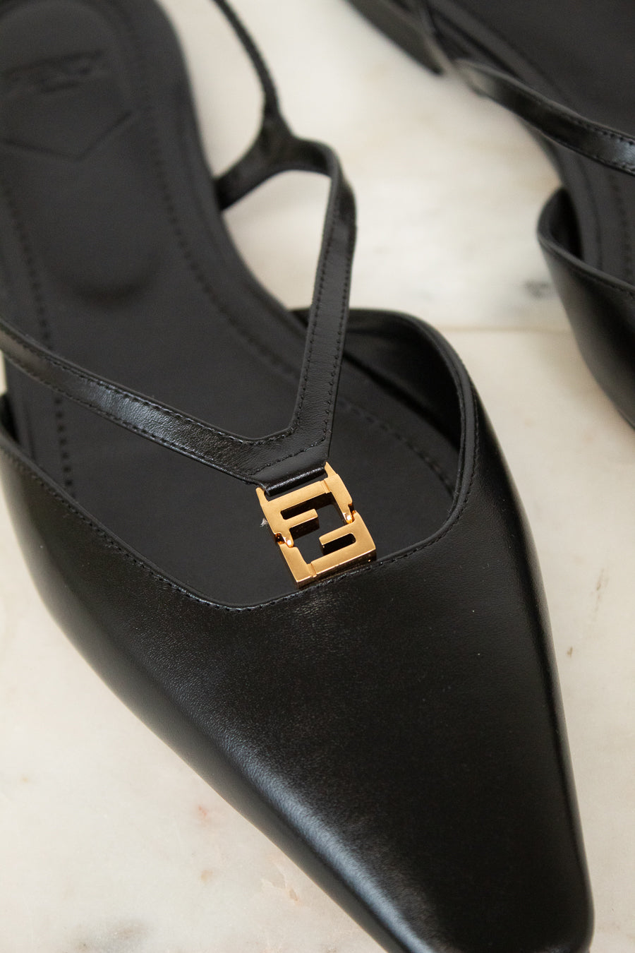 A close-up of a black leather pointed-toe slingback shoe with a gold-tone interlocking FF logo hardware on the strap, resting on a light marble surface.