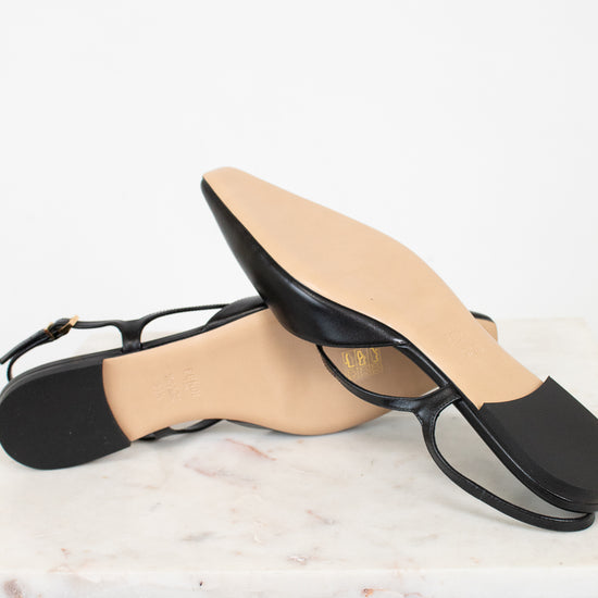 A pair of black leather flat sandals with square toes and thin straps, resting on a white marble slab against a white background.