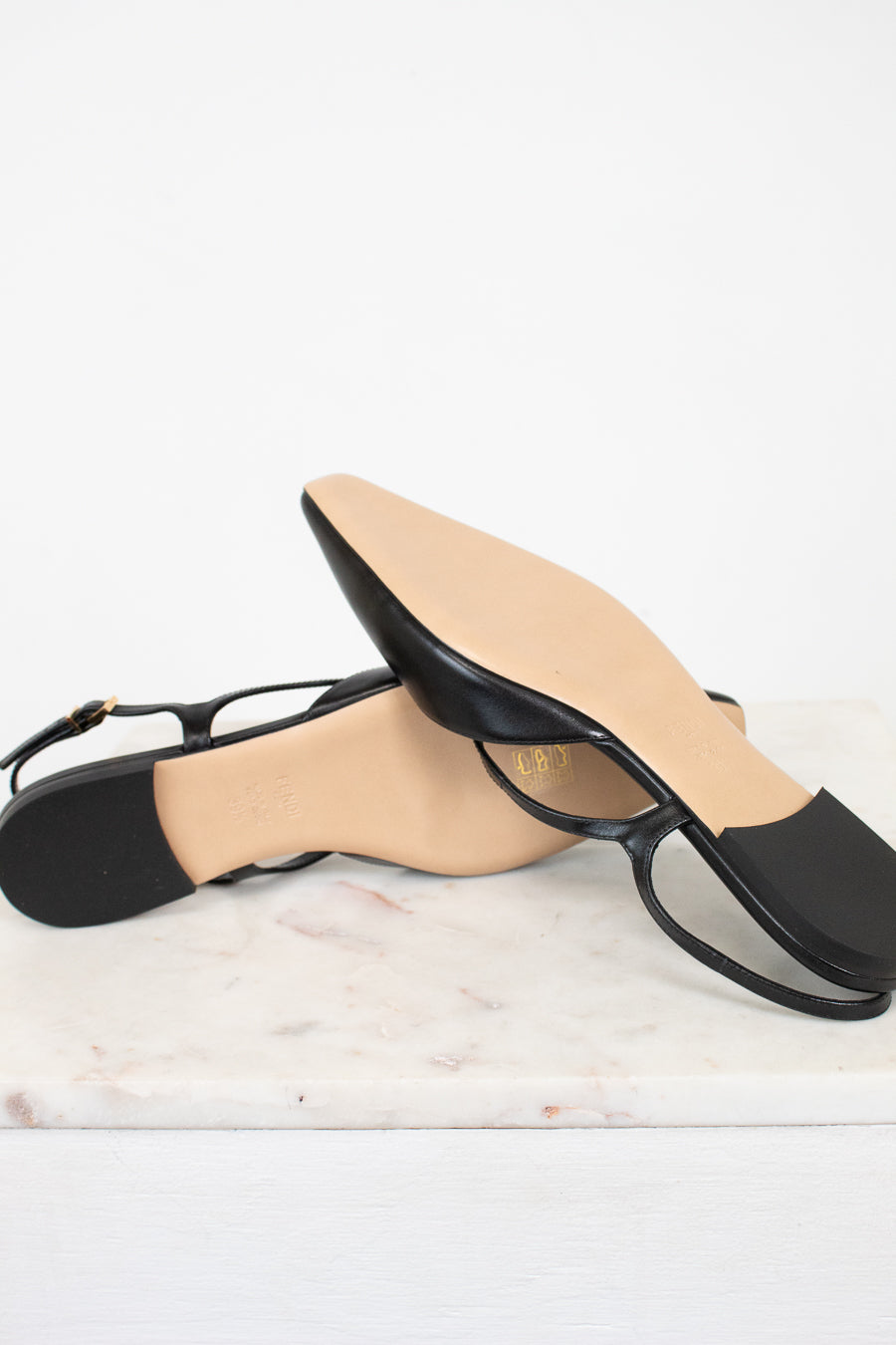 A pair of black leather flat sandals with square toes and thin straps, resting on a white marble slab against a white background.