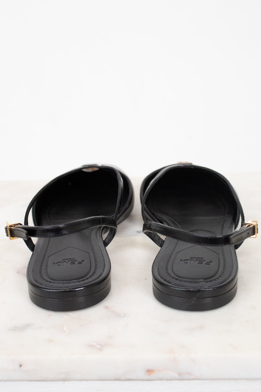 A pair of black Fendi slingback flat sandals with gold-tone buckle details, shown from the back on a white marble surface against a white background.
