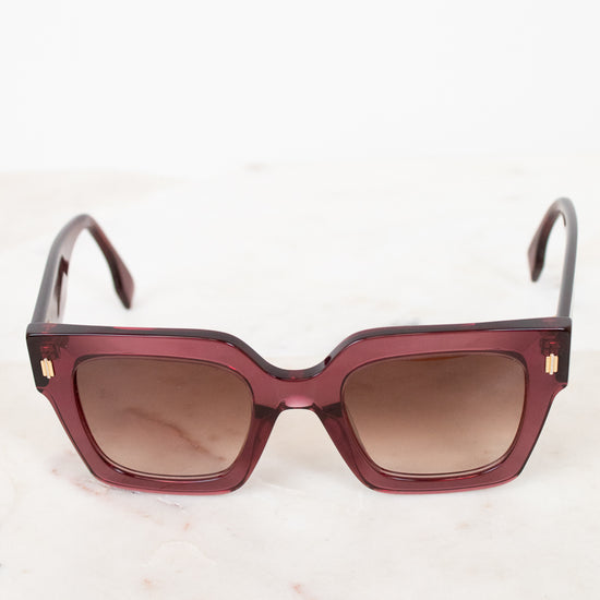 Sunglasses with a purple tinted frame and brown gradient lenses on a light background