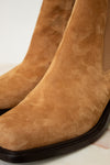Close-up of a tan suede ankle boot with a dark sole, showing the side elastic gore and textured suede upper.