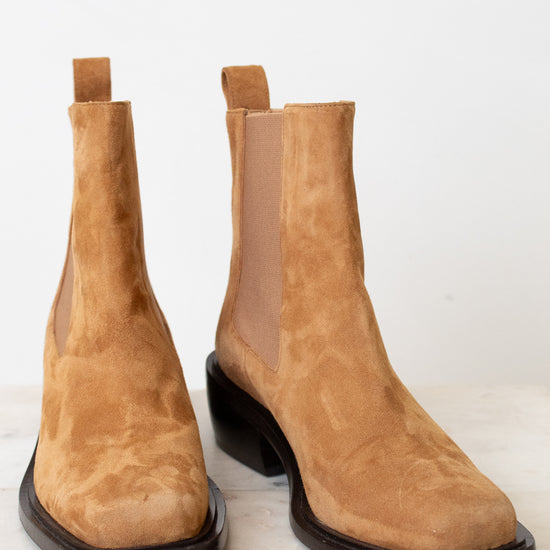 A pair of tan suede Chelsea boots with dark brown square soles, displayed on a light marble surface against a white background.