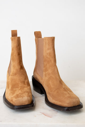 A pair of tan suede Chelsea boots with dark brown square soles, displayed on a light marble surface against a white background.