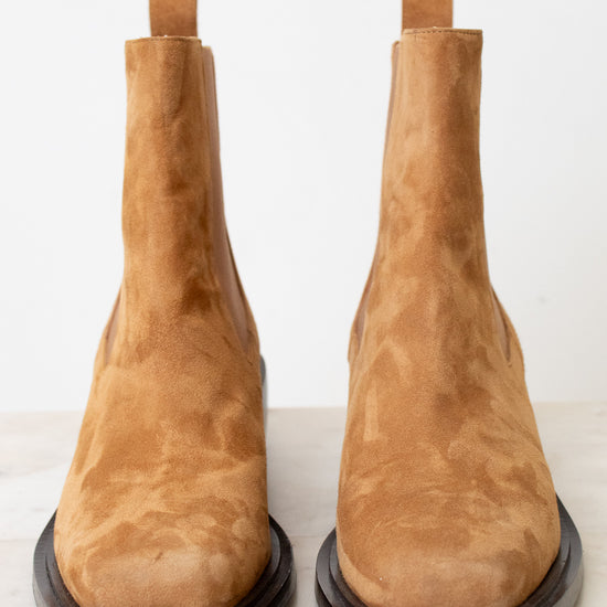 A pair of tan suede Chelsea boots with dark brown soles, photographed from the front on a light marble surface against a white background.