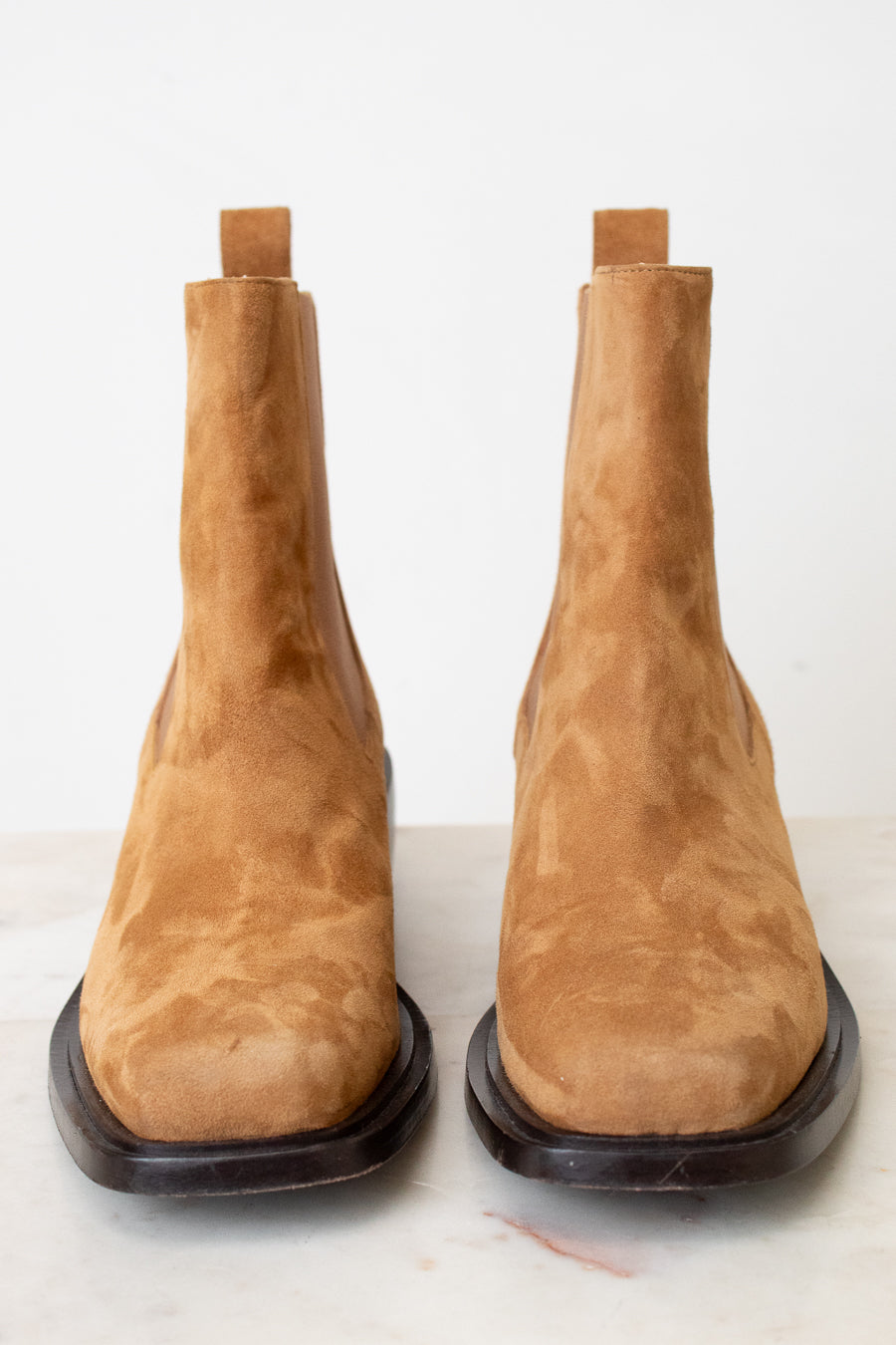 A pair of tan suede Chelsea boots with dark brown soles, photographed from the front on a light marble surface against a white background.