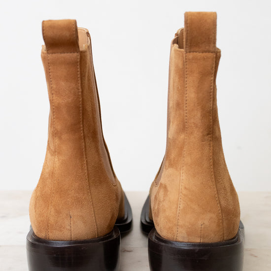 A pair of tan suede Chelsea-style ankle boots with black soles, shown from the back on a light marble surface against a white background.