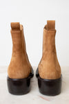 A pair of tan suede Chelsea-style ankle boots with black soles, shown from the back on a light marble surface against a white background.