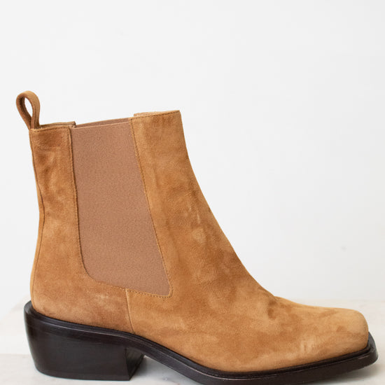 A tan suede Chelsea boot with a brown elastic side panel and a dark brown block heel, shown from a side profile on a light marble surface against a white background.
