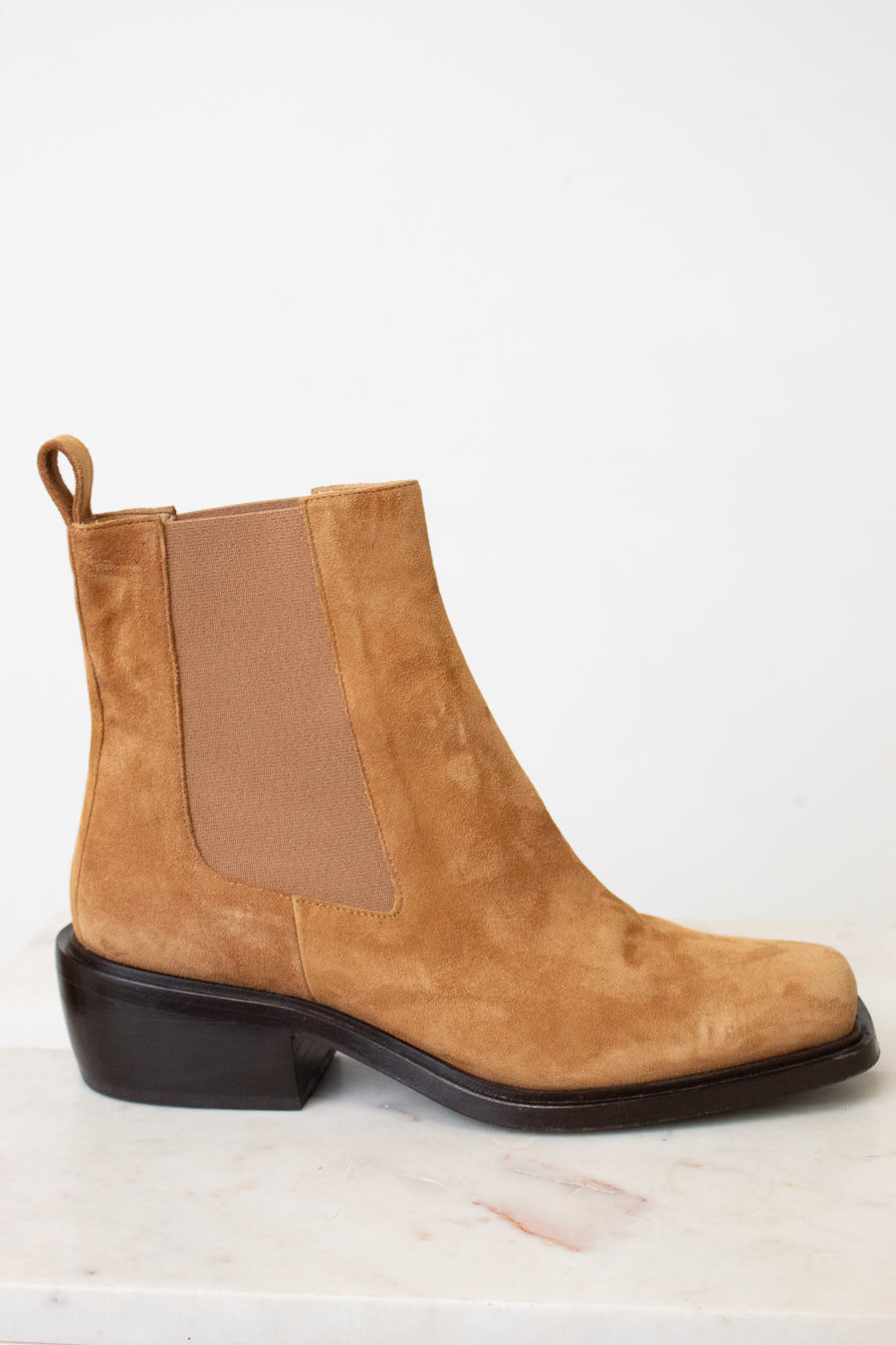 A tan suede Chelsea boot with a brown elastic side panel and a dark brown block heel, shown from a side profile on a light marble surface against a white background.
