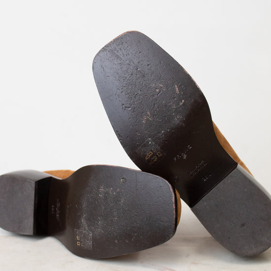 Close-up of the soles of a pair of brown leather shoes resting on a light marble surface against a white background.