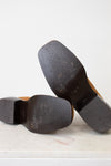Close-up of the soles of a pair of brown leather shoes resting on a light marble surface against a white background.