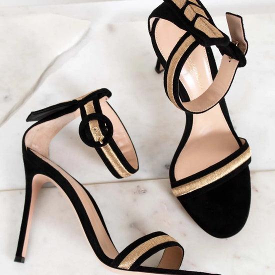 Black high-heeled sandals with gold strap detail on a light background