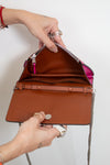 A brown leather interior of hot pink clutch handbag with a silver chain strap, held open by a person's hands to show the interior compartments against a plain white background.
