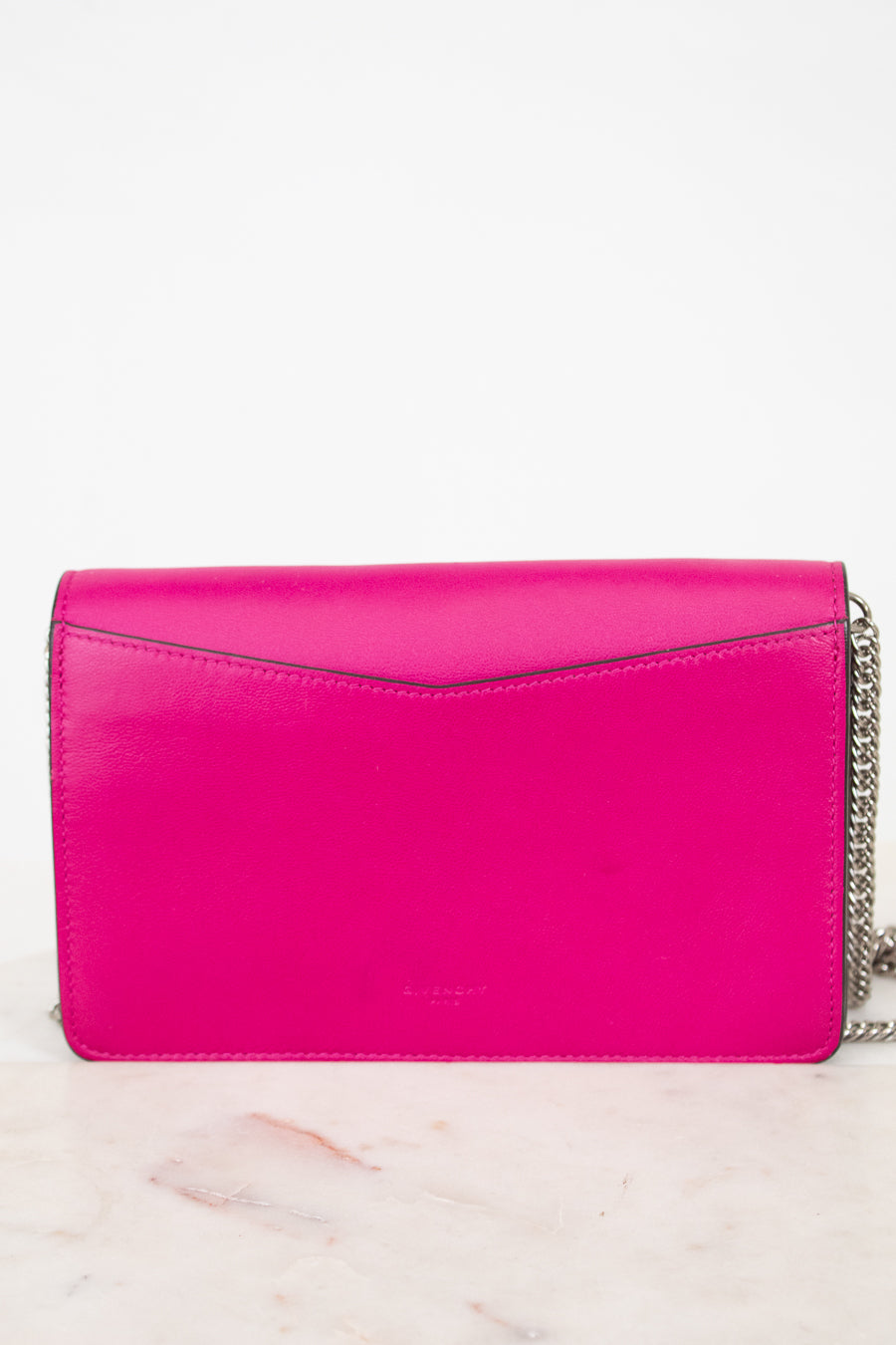 A bright hot pink leather clutch bag with a silver chain strap, resting on a light marble surface against a white background.