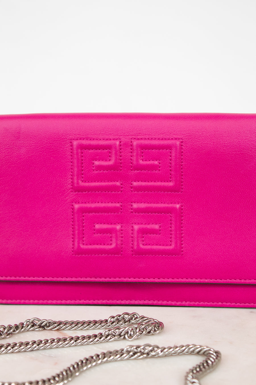 A close-up of a bright pink Givenchy envelope clutch bag with the brand's signature embossed logo on the front flap and a silver chain strap resting below it.
