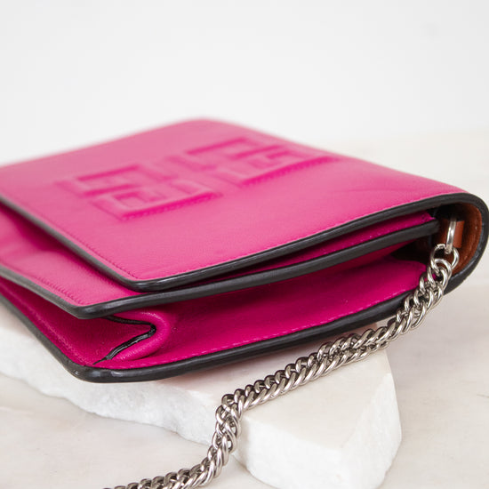 A bright pink leather crossbody bag with a silver chain strap, resting on a white marble block against a white background.