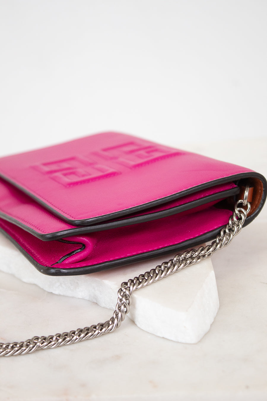 A bright pink leather crossbody bag with a silver chain strap, resting on a white marble block against a white background.