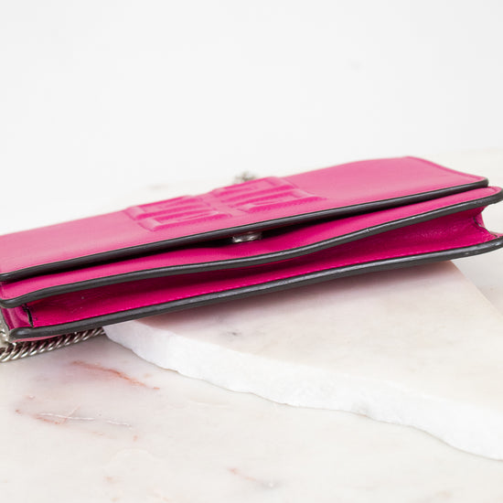 A bright pink leather clutch wallet with a silver chain strap, resting on a white marble slab against a white background.