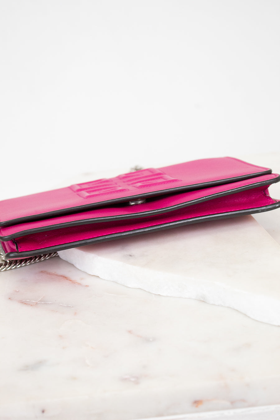 A bright pink leather clutch wallet with a silver chain strap, resting on a white marble slab against a white background.