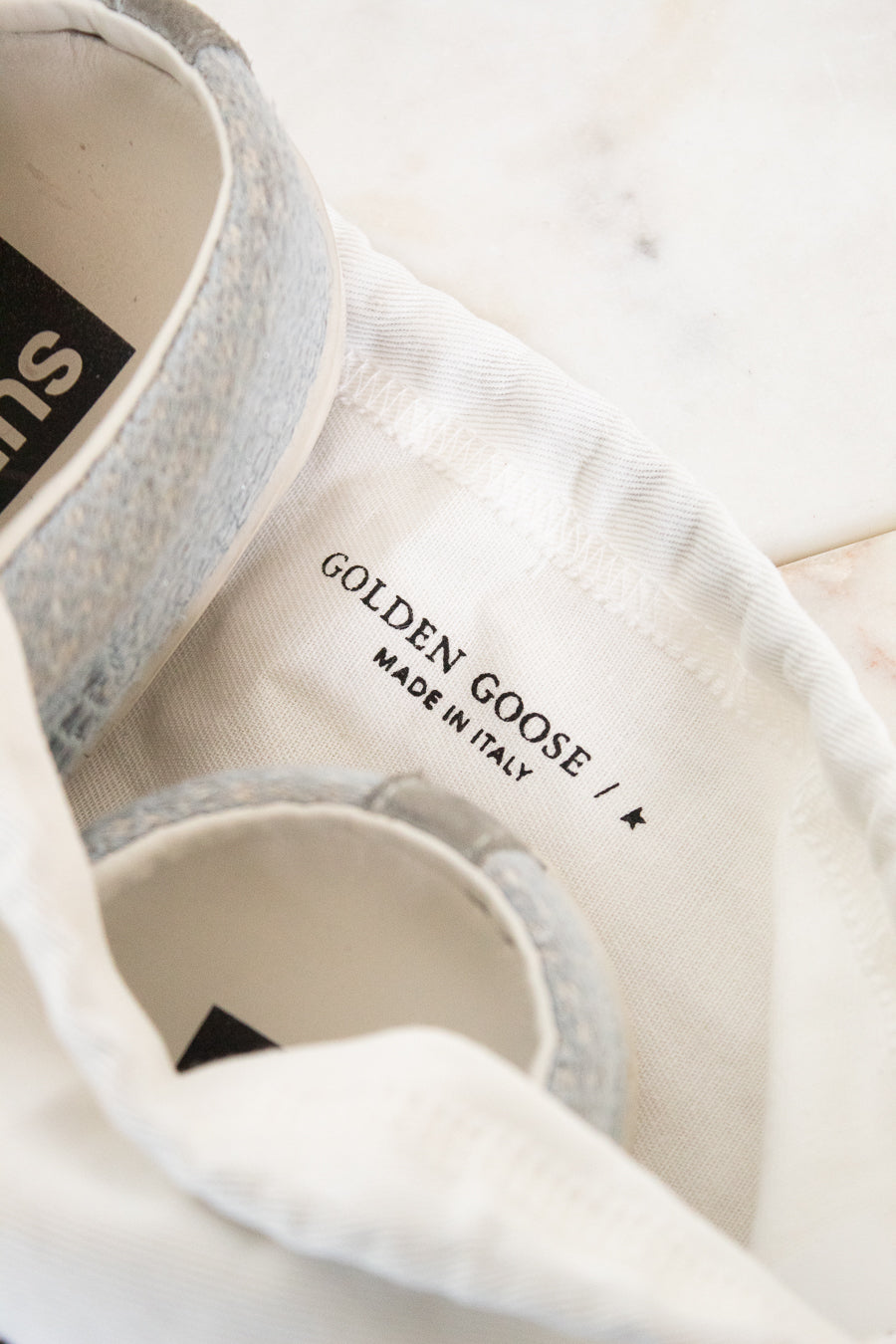 Close-up of a white canvas shoe bag with a light gray fabric shoe partially tucked inside. The interior label reads 'GOLDEN GOOSE / MADE IN ITALY'.