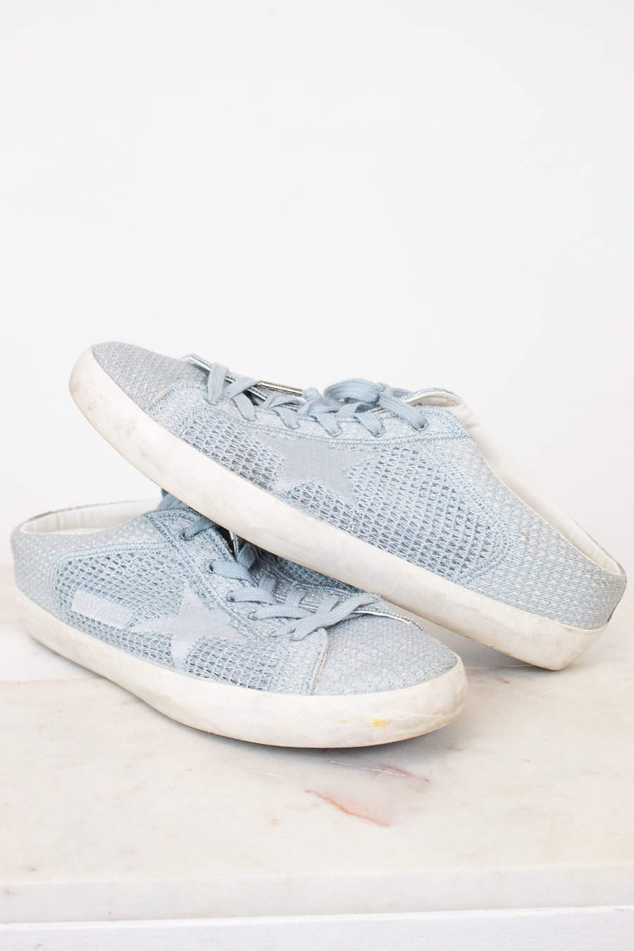 A pair of light blue mesh sneakers with white rubber soles, displayed on a light marble surface against a white background.