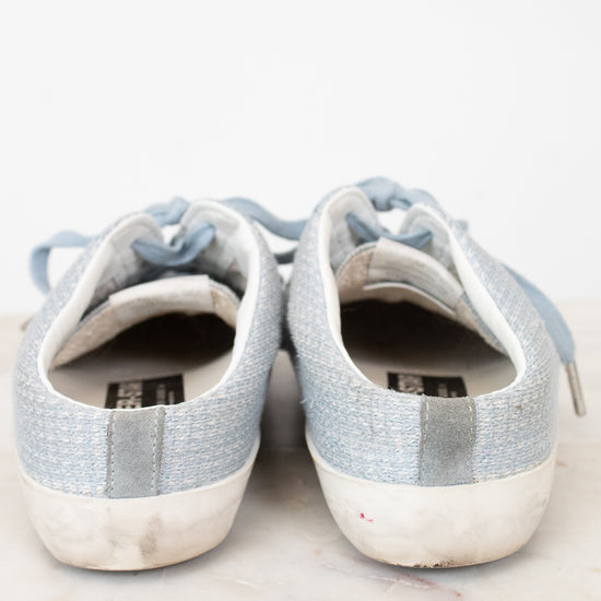 A pair of light blue canvas low-top sneakers with white rubber soles, shown from the back on a light marble surface against a white background.