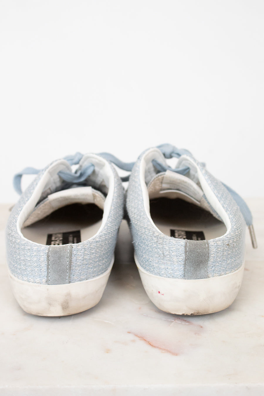 A pair of light blue canvas low-top sneakers with white rubber soles, shown from the back on a light marble surface against a white background.