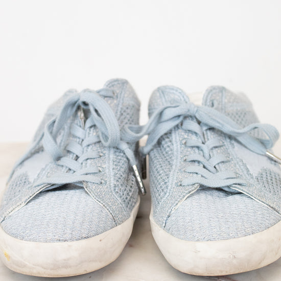 A pair of light blue denim sneakers with white rubber soles, shown from the front on a light marble surface against a white background.