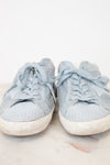 A pair of light blue denim sneakers with white rubber soles, shown from the front on a light marble surface against a white background.