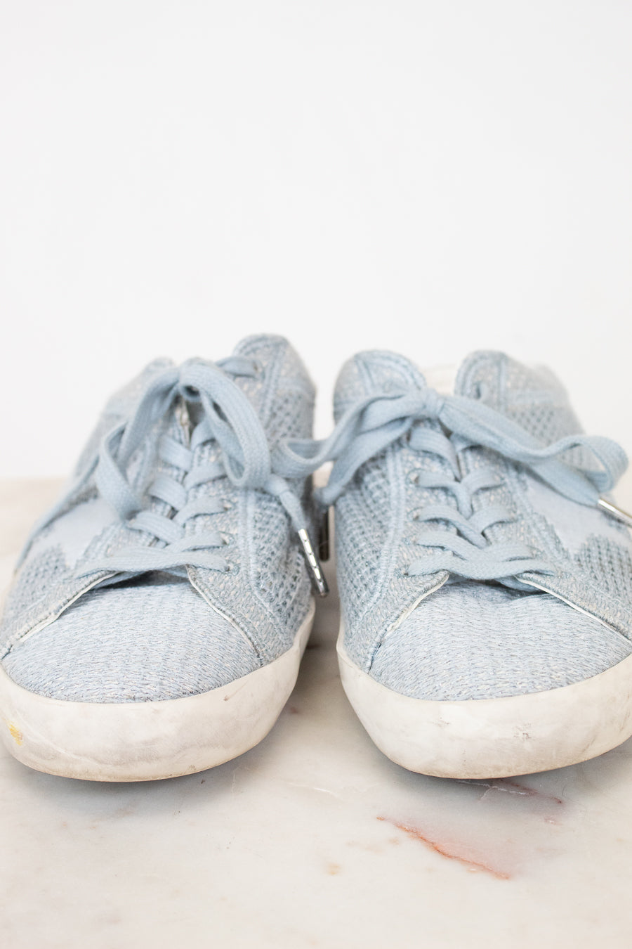 A pair of light blue denim sneakers with white rubber soles, shown from the front on a light marble surface against a white background.