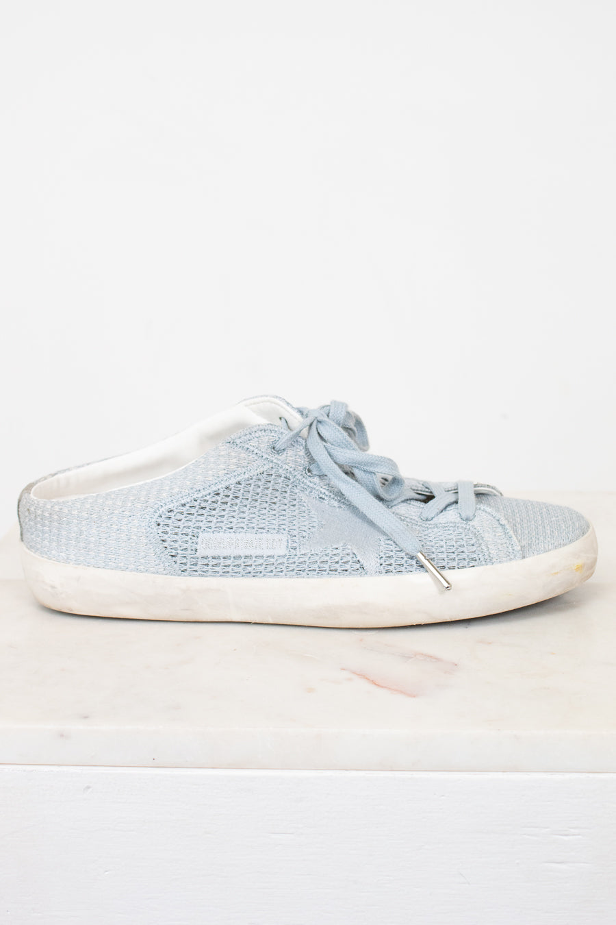 A light blue textured slip-on sneaker with matching laces and a distressed off-white sole, displayed on a white marble surface against a white background.