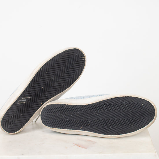 A pair of light blue canvas slip-on shoes shown from the bottom, displaying their black chevron-patterned rubber soles with white edges, set against a plain white background.