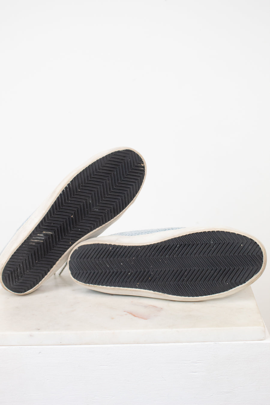 A pair of light blue canvas slip-on shoes shown from the bottom, displaying their black chevron-patterned rubber soles with white edges, set against a plain white background.