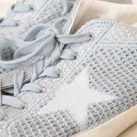 A close-up of a light blue knit sneaker with matching blue laces and a white star detail on the side, shown against a neutral cream background.