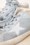 A close-up of a light blue knit sneaker with matching blue laces and a white star detail on the side, shown against a neutral cream background.