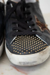 Black sneaker with gold studs on a white surface