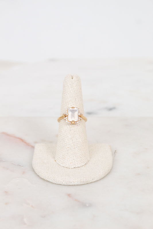 Gold ring with a moon quartz gemstone on a textured white stand against a light marble background