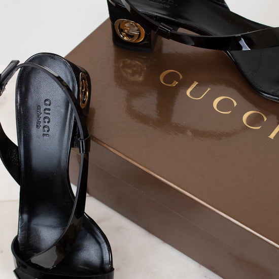 Black Gucci high-heeled sandals on a white surface with a Gucci box in the background.