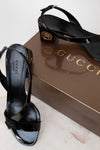 Black Gucci high-heeled sandals on a white surface with a Gucci box in the background.