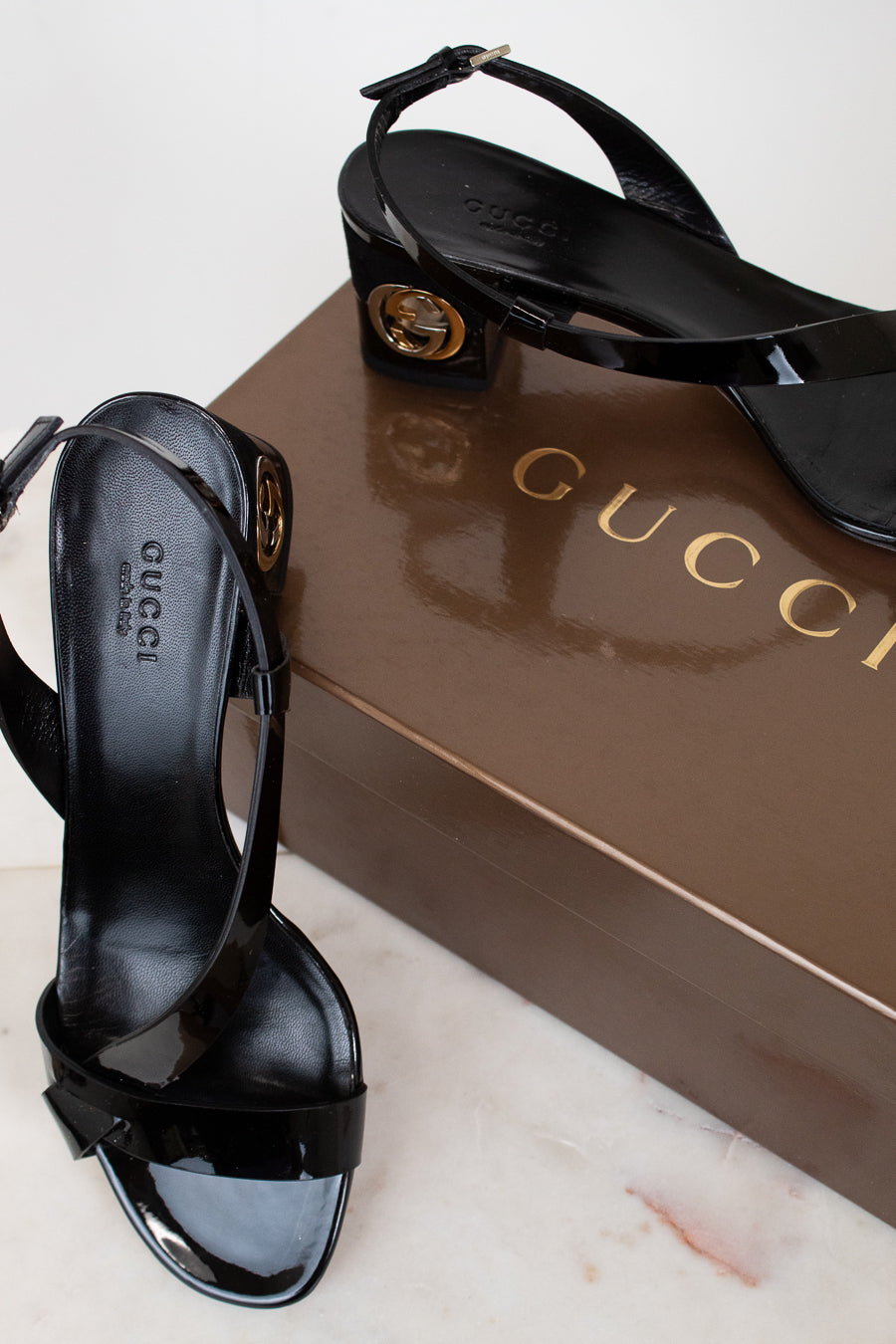 Black Gucci high-heeled sandals on a white surface with a Gucci box in the background.