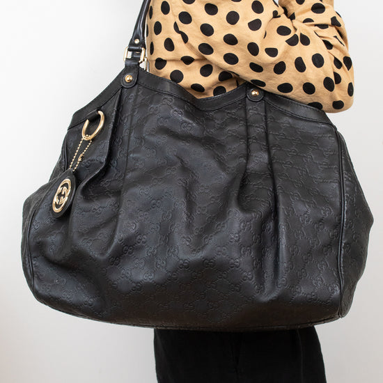 A person wearing a beige and black polka-dot top carries a large black leather shoulder bag with gold-tone hardware and a circular interlocking G logo charm. The person is also wearing black pants and gold hoop earrings, photographed against a plain light gray background.
