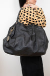 A person wearing a beige and black polka-dot top carries a large black leather shoulder bag with gold-tone hardware and a circular interlocking G logo charm. The person is also wearing black pants and gold hoop earrings, photographed against a plain light gray background.